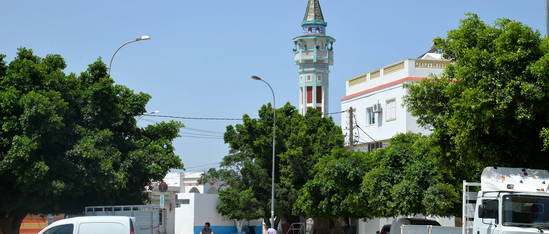 Vue d’une avenue à Aousja avec des bâtiments, des arbres et un minaret au loin